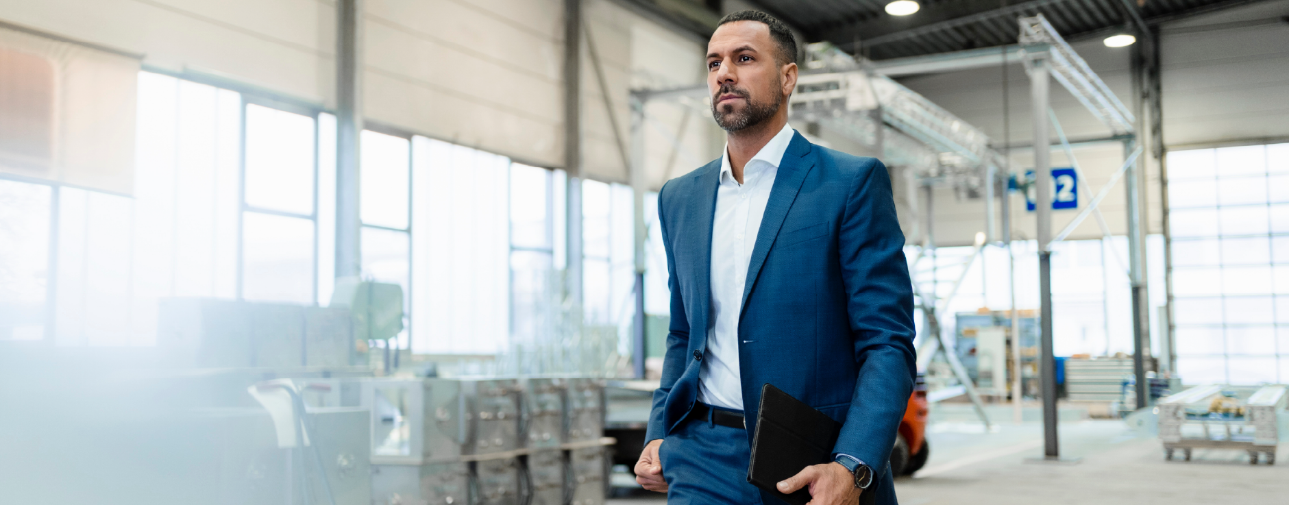 homem de barba vestindo um terno azul caminhando em um galpo de empresa carregando um tablet